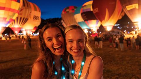 Two women hug and smile at the camera with hot air balloons in the background