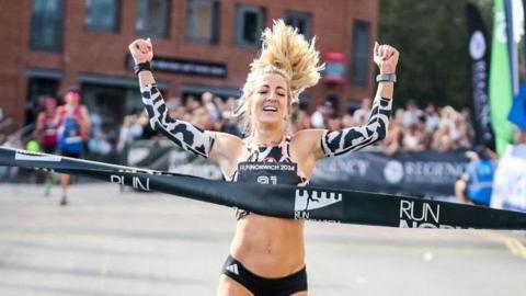 Holly Archer, who has blonde hair and is wearing black running pants and black and white arm warmers, crossing the finish line with spectators in the background. She is holding her arms above her head as she celebrates.