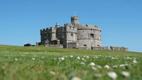 Pendennis Castle stands on a grassy hill dotted with small white flowers, with the stone keep and fortifications set against a clear blue sky.