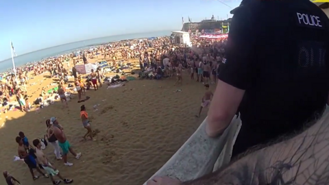 A large mob of young people, mainly men and boys in beach wear, stood on a sunny and busy beach. The picture is taken from a high vantage point with a police bodycam. Another police officer is standing at the railing.