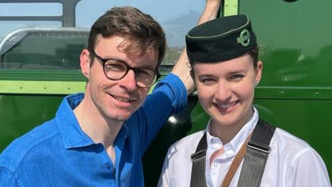 A man with short brown hair and a blue shirt is stood with a woman dressed as a bus conductor with a short green hat and white shirt.