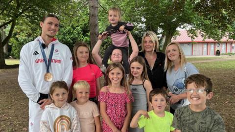 Lewis Davey wearing his white Great Britain tracksuit with his bronze medal around his neck stands in a woodland area, posing for a photo with women and children. Davey has dark hair with a parting down the middle.