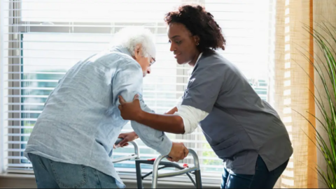An elderly man in a shirt and jeans is helped up out of a chair by a nurse. The man is holding a walking frame and is positioned by a widow. The nurse has a grey uniform top on and dark curly hair.
