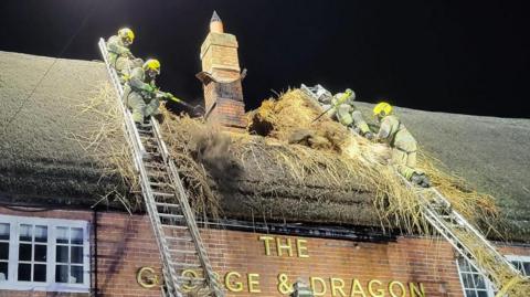 Four firefighters standing at the top of silver ladders, leaning up against the thatched roof of the George and Dragon pub. It is a red brick building with sash windows and a tall chimney in the middle. The sky is dark and the fire is extinguished, but the thatched roof has been torn and burnt.