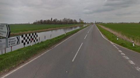 A long, straight rural road runs alongside a water-filled ditch. There are road markings for a junction to the right and road signs to the left.