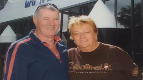 A man in a polo shirt and a woman in a t-shirt. Both are looking at the camera and smiling. They have an arm around each other.
