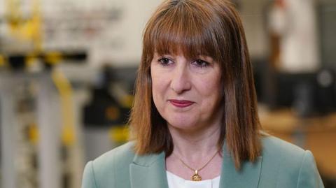 Chancellor Rachel Reeves wearing a light green jacket and white top standing in front of a blurred background