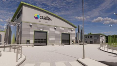 A computer generated image of proposed anaerobic digestion site showing grey building with a slanted green roof surrounded on two sides by large metal cylinders 