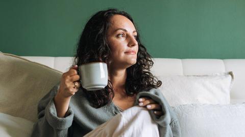 A woman looking into the distance while sat on a bed holding a cup