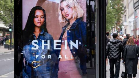 A bus stop advertising screen displays an add for Shein featuring two women, one wearing a denim jumpsuit and the other wearing pink activewear and a denim jacket.