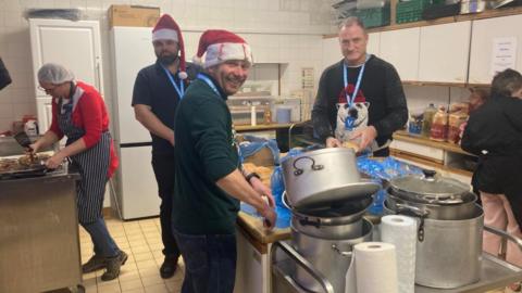 Three men in a kitchen preparing food. Two of them are wearing Christmas hats. A woman is to the left preparing food on a different worktop, as well as another woman on the right. There are metal pots and pans piled up in front of the worktop in the middle.