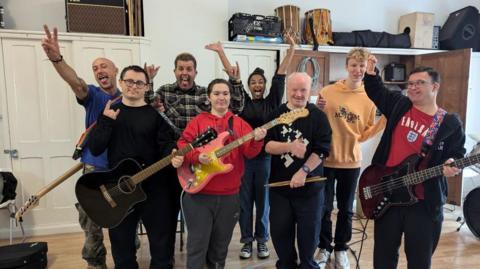 Delta Sonic band members with charity volunteers. Most are holding instruments, including guitars and one man is holding drumsticks. 