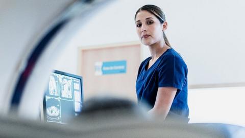 A female healthcare professional looks over as a patient is moved into a CT scanner, the patient is out of focus and the image is taken from inside the scanner itself, with a screen showing scans and a white hospital room in the background.