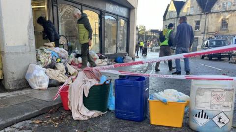 People clearing out a shop in Bradford-on-Avon following flooding. Blankets and towels can be seen on the pavement outside, as well as several buckets. There is also red and white tape across part of the road, that says do not cross. 