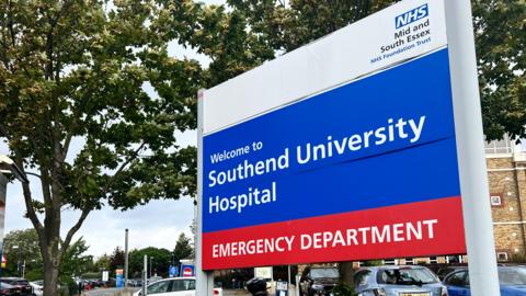 General view of Southend Hospital's entrance. It shows a large sign detailing the hospital name and the nearby emergency department in large white letters against a blue and red background. 