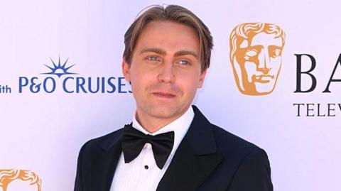 Barney poses on a red carpet, in front of a white backdrop with with Bafta branding on it. He has dark blonde hair, and wears a black tuxedo with a black bow tie and a white shirt. 