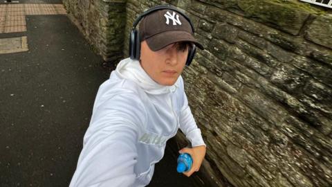 Finlay Thornell. He is out running, wearing a white hoodie, a brown cap and black headphones. He is holding a blue bottle. He is looking at the camera, although his face is partially covered by the hat.