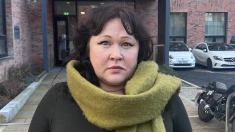 Ms Morrison has dark black hair and is wearing makeup including a winged eyeliner. She has a bright green scarf and a black top.