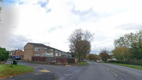 A junction. There are houses on the left side with trees and cars driving across the road 