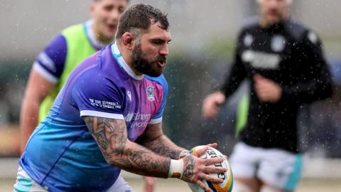 Jamie Bhatti in the purple shirt of Glasgow Warriors holds the ball during a warm-up, with two team-mates indistinct in the background.