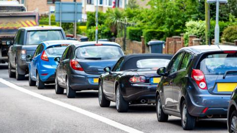 A queue of cars, all of different shapes and sized, queued up on a residential street. 