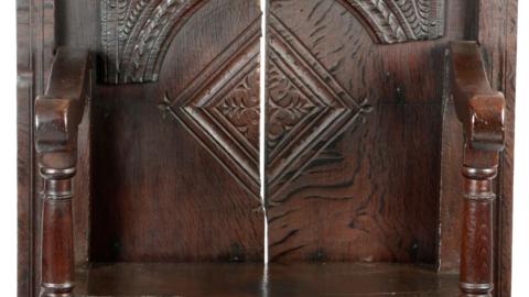 A close-up image of a dark wooden chair. The chair has a noticeable gap in the middle of its back and is carved with elaborate illustrations.
