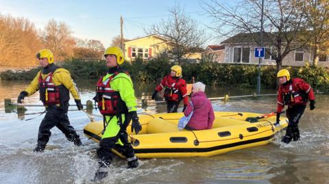 Four firefighters guiding a yellow inflatable boat, with a woman on board wearing a purple anorak, through floodwater. There are mobile homes in the background.