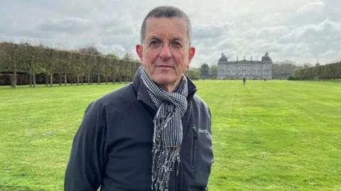 Antony Gormley is standing looking at the camera, wearing a dark grey/navy outdoor jacket and a scarf. In the distance behind him there is a large stately home.