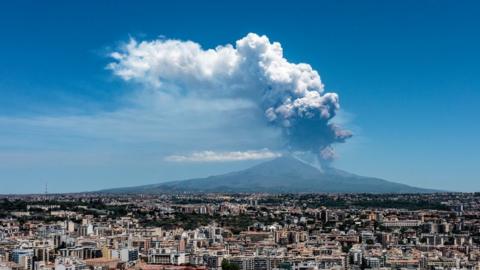 Dark grey and white plumes are seen rising from Mount Etna with the rooftops of buildings in the foreground