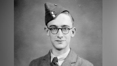 Young man wearing glasses and a military uniform. He is looking into the camera with a serious expression and wearing his RAF hat tilted to the side of his head.
