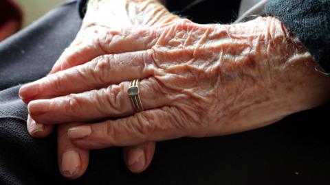 File image showing an elderly woman, wearing chipped pale pink nail polish and a gold ring with a blue stone, crossing her hands on her lap