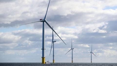 Four of five new wind turbines at the Blyth offshore wind farm. There are four large wind turbines that are silver and have yellow bollards at the bottom. There is a ship near the first wind wind turbine. 