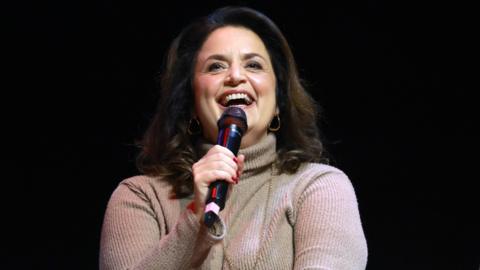 Ruth Jones talks into a black microphone. She is smiling and wears a cream coloured jumper. She has shoulder length brown hair and is wearing gold hoop earrings and a gold necklace.