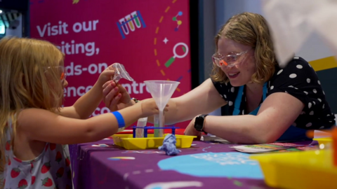 A side-on view of a girl aged about 8 with long fair hair hiding her face and wearing safety goggles, reaching out across a table to a woman who is handing her a small plastic bag. The woman has chin-length light brown hair and is wearing a black top with white dotgs and safety goggles. She is leaning over the table and on it is resting various bits of brightly coloured plastic, including a clear funnel. 