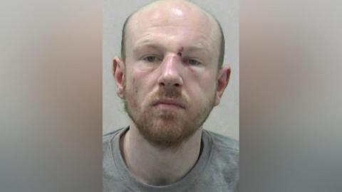 Mugshot of Armstrong. He has a short ginger beard and a bald head, with a cut around his left eye.