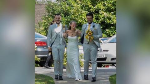 Melanie Waterhouse walks towards the camera in her wedding dress and supported by her two sons in morning suits