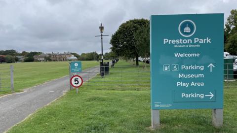 An upright blue sign at the entrance to Preston Park Museum & Grounds. A grassed area with trees can be seen in the background. A 5mph speed limit sign is positioned next to the driveway.