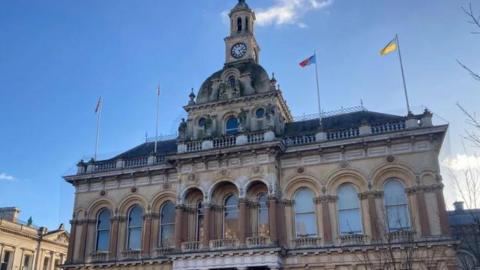 The upper floor of Ipswich Town Hall pictured from Cornhill, with the clock on top of the central dome and four flags on flagpoles on the front of the roof