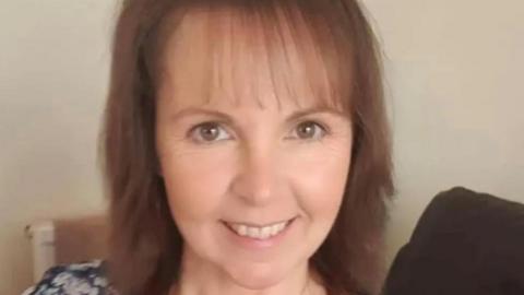 Heather Smedley, who has brown shoulder length hair, smiles while sat on a sofa.