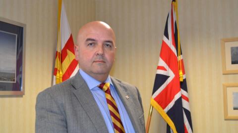 Jonathan Le Tocq stood in a jacket and tie in front of two flags