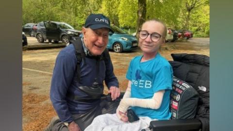 A man knelt next to a girl in a wheelchair wearing a blue t-shirt.