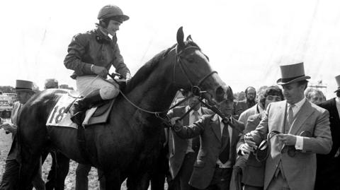 
It is a black and white image of a smiling jockey sitting on top of a racehorse which is wearing a saddle with the number eighteen on it. Another man dressed in a top hat and morning suit is standing beside the horse holding the reigns. There is a crowd of men standing behind the men wearing the top hat.