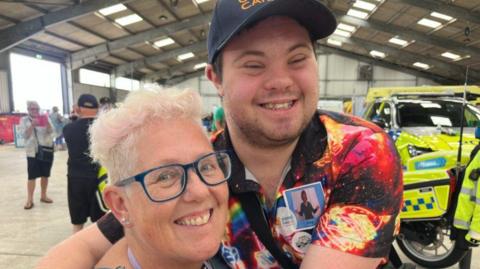 Mum Jenny and her son Charlie smiling at the camera. Charlie is hugging his mum. There are police vehicles in the background.