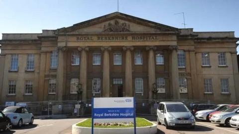 A large stone building with five tall pillars out the front, behind a car park with a blue and white sign saying 'Royal Berkshire Hospital'