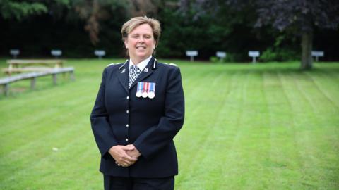 Debbie Tedds standing in front of green grass. She is wearing a navy police suit with a black and white checkerboard tie, and has three medals on her chest.