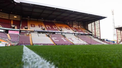 An empty football stadium, photographed from one of the pitch's touchlines. The words 'Bradford City' are visible across the seats in the stand visible.