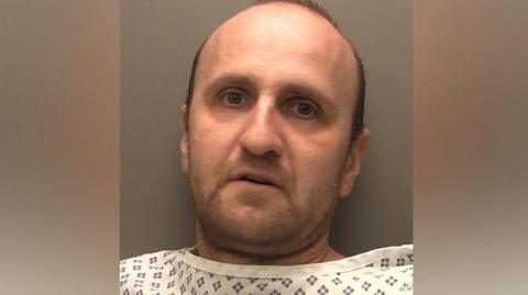 Custody photograph of Veselin Dudenski. He has short black hair and beard and is wearing a white top with black squares on.