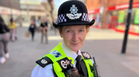 Woman with black hat and high-vis jacket. She is stood in a street smiling softly at the camera