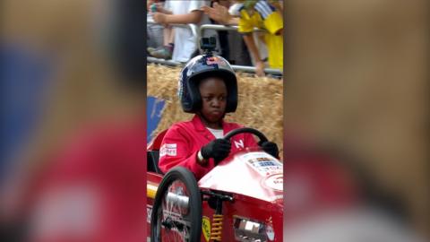 A young woman in red overalls and a crash helmet sits behind the steering wheel of a soapbox.