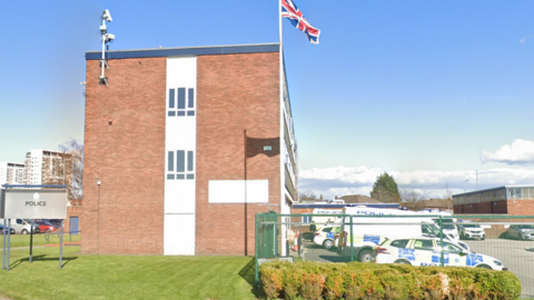 Kirkby police station in Kirkby, Merseyside. It is a three-storey brick building with a number of police cars and vans parked in its car park.